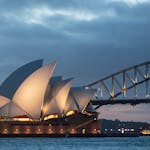 Facade of modern Sydney Opera House of unusual design with large concrete shells located on river coast near massive bridge at night